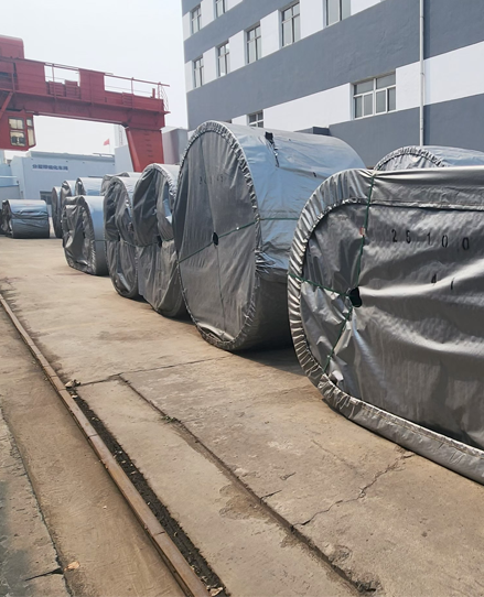 Outdoor factory storage area with a red gantry crane and massive, heavy-duty conveyor belt rolls packaged for global export.