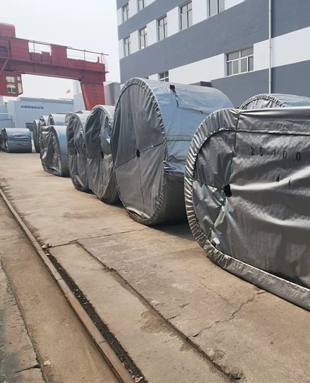 Outdoor factory storage area with a red gantry crane and massive, heavy-duty conveyor belt rolls packaged for global export.