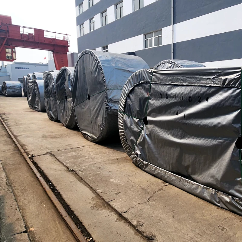 Outdoor factory storage area with a red gantry crane and massive, heavy-duty conveyor belt rolls packaged for global export.